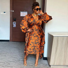 Load image into Gallery viewer, Woman in an orange and black patterned dress standing indoors next to a door.
